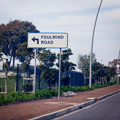 Image showing Signage for wind, road and city with information, outdoor and nature with urban town and design. Direction, icon and street with symbol and trees with poster and language with bushes and board