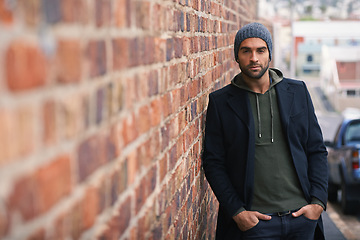 Image showing Portrait, confidence and man on brick wall in city in casual clothes, outfit or fashion for winter outdoor. Serious, cool and stylish person in urban town in beanie, hoodie and trendy jacket in Spain
