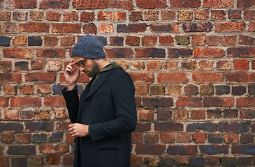 Image showing Fashion, man and thinking on brick wall with glasses for trendy, cool and style for urban with idea. Male person, outdoor and planning for weekend or holiday with winter outfit and stylish in Germany