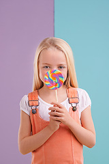 Image showing Studio, girl and hiding with lollipop, dessert and playful for snack and childhood. Child, sweets and yummy candy for tasty, eating and happiness on vibrant split pastel pink and blue background