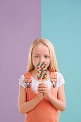 Image showing Studio, child and hiding with lollipop, desert and playful for snack and childhood. Girl, sweets and yummy candy for tasty, eating and happiness on vibrant split pastel pink and blue background