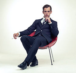 Image showing Suit, smoking and man portrait in chair, studio and serious in formal fashion by white background. Young model, face and elegant clothes with cigarette in armchair and confident in tuxedo by party