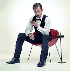 Image showing Smoking, tuxedo and portrait of man in studio with red chair and cigarette for elegant and fancy outfit. Serious, handsome and male model with bowtie and suit for classy fashion by white background.