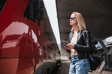 Image showing Young blond woman in jeans, shirt and leather jacket wearing bag and sunglass, embarking red modern speed train on train station platform. Travel and transportation.