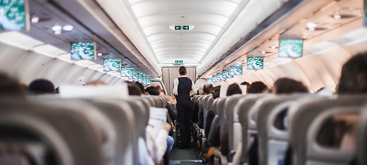Image showing Interior of airplane with passengers on seats and stewardess in uniform walking the aisle, serving people. Commercial economy flight service concept.