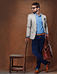 Image showing Bag, chair and man with suit for fashion, style and formal wear isolated on studio background. Male person, gentleman and businessman with trendy clothes, class and outfit in brown backdrop