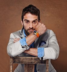 Image showing Fashion, watch and portrait of man in studio with trendy, stylish and classy suit on chair. Serious, time and handsome male person with elegant style for outfit and accessory by brown background.