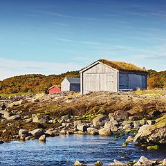 Image showing Norway, house and outdoor in field, lake and rocks for travel and holiday in background with hills. Shed, grass and blue sky for cottage, serenity and sunlight for nature in season for countryside