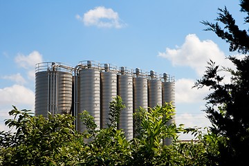 Image showing Twelve high metal tower silos on chemical plant behind the trees