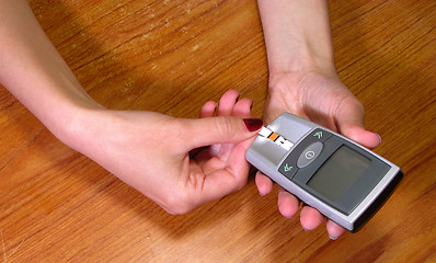 Image showing Two hands introducing a glicemic test strip in the glucometer on a wooden desk-educational image for diabetes patients.