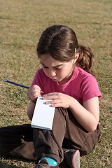 Image showing Cute little girl with pigtails writes in the notepad sitting on the grass