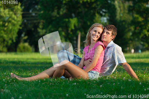 Image of Two young lovers sitting on the lawn in the park and looks into 