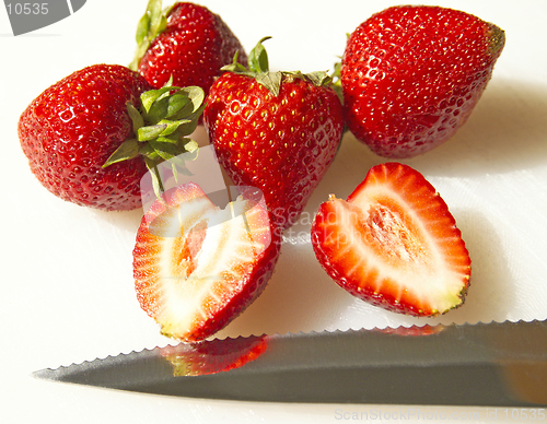 Image of A sunlit sliced strawberry rests with other whole strawberries with a knife on a white cutting board (macro-14MP camera)