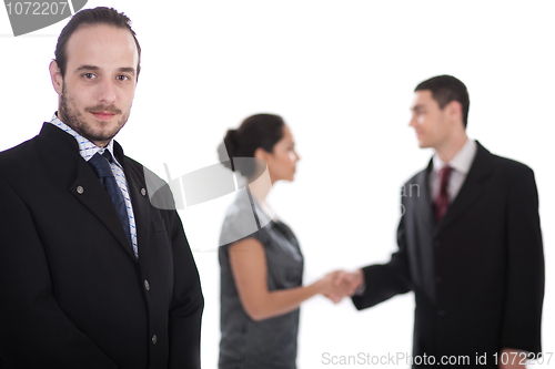 Image of Handsome business man looking at us, two collegues giving shakehand at the background