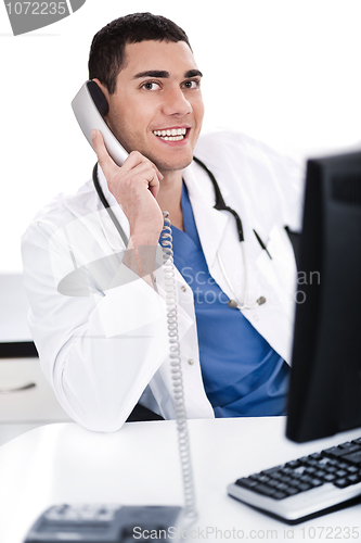 Image of Smiling young physician sitting at his desk talking over phone