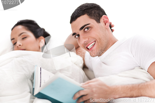 Image of Woman sleeping and man happily reading book in bed