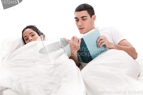 Image of Young handsome man reading book while is wife sleeping beside him in bed