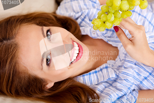 Image of Happy young woman lying on sofa holding grapes