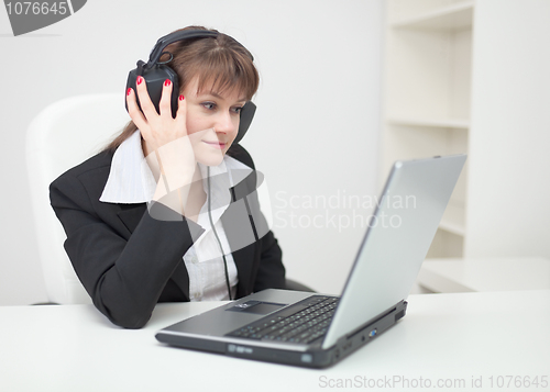 Image of Woman in ear-phones sits with laptop at table at office