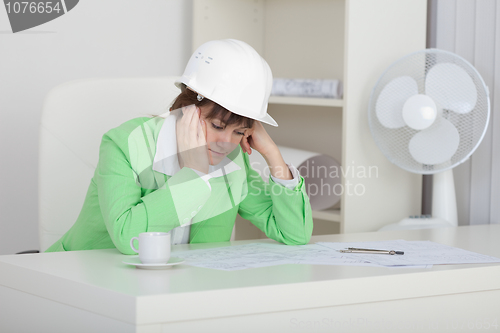 Image of Sad woman engineer with helmet on head sits at table on workplac