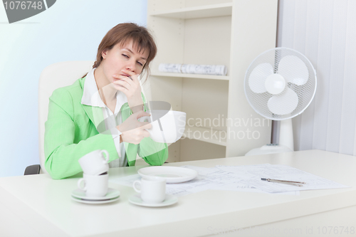 Image of Tired woman sits at table with big cup of coffee and yawns