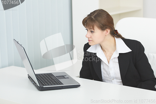 Image of Young serious woman sits at table at light office with laptop