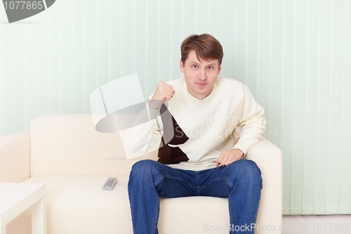 Image of Young man sits on sofa and watches football on TV