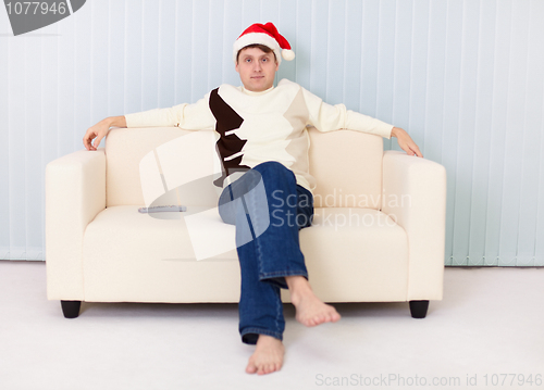 Image of Young man in Christmas hat sits on sofa and watches TV