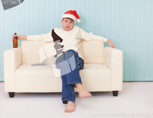 Image of Man in a New Year's cap sits on sofa with bottle