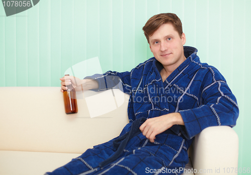 Image of Young man drinks beer in dressing gown on sofa