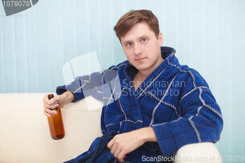 Image of Young man in dressing gown sits on a couch with bottle