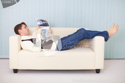 Image of Young man lies comfortably on sofa with a newspaper