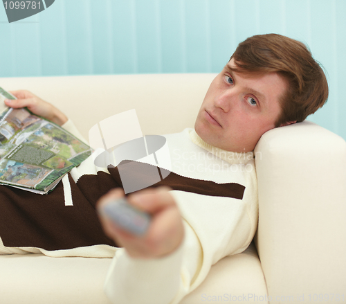 Image of Young man, lying on couch with magazine and TV remote control