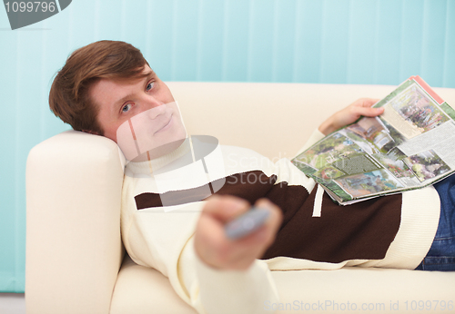 Image of Smiling man, lying on couch with magazine and TV remote control
