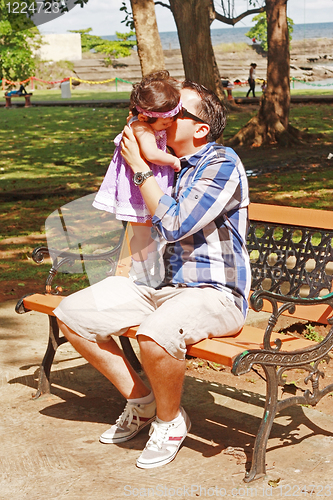 Image of A father holds and kisses his baby with a muted tropical setting in the background