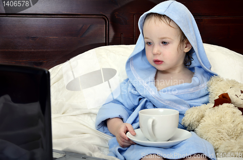 Image of little girl in a bathrobe relaxing on the bed after a shower with cup of tea and laptop