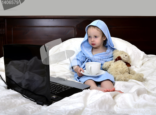 Image of little girl in a bathrobe relaxing on the bed after a shower with cup of tea and laptop