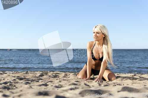 Image of blonde girl in a black bathing suit sitting on shore blue sea 