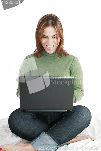 Image of Excited girl in jeans with laptop sitting on her bed isolated 