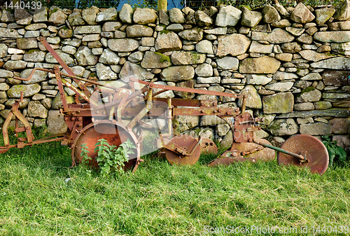 Image of Old rusty plow in shadow of stone wall