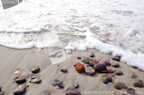 Image of Sea waves beat stones lying in sand on coast line.