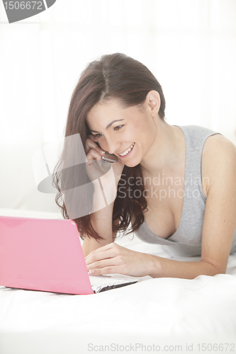 Image of Girl smiles listening to conversation  on the phone while using computer  in bed