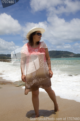 Image of A woman in a white hat and a pink dress walking on the beach