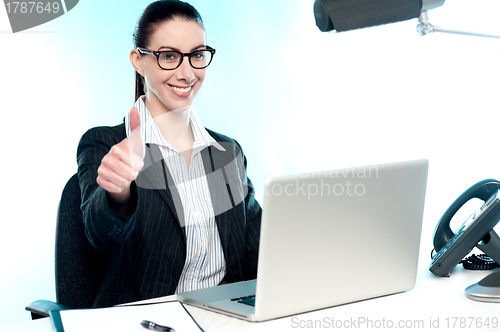 Image of Young businesswoman sitting on desk  gesturing thumbs up