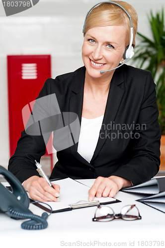 Image of Smiling woman wearing headset writing on notepad