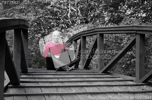 Image of young businessman in red shirt working on laptop at bridge