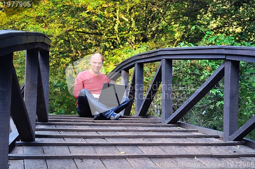 Image of young businessman in red shirt at bridge  working on laptop 