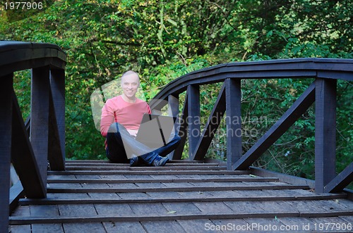 Image of young businessman in red shirt at bridge  working on laptop 