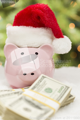 Image of Pink Piggy Bank Wearing Santa Hat Near Stacks of Money on Snowfl