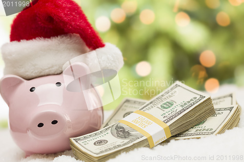 Image of Pink Piggy Bank Wearing Santa Hat Near Stacks of Money on Snowfl